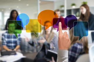 Eine Gruppe von Menschen sitzt in einem modernen Besprechungsraum und diskutiert. Im Vordergrund zeigt eine Hand auf eine Reihe bunter, transparenter Sprechblasen, die digital über das Bild gelegt sind. Die Szene symbolisiert Kommunikation, Diskussion und Beteiligung