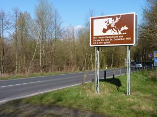 Straße mit Schild zur Erinnerung. Im Hintergrund stehen Bäume. 