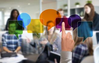 Eine Gruppe von Menschen sitzt in einem modernen Besprechungsraum und diskutiert. Im Vordergrund zeigt eine Hand auf eine Reihe bunter, transparenter Sprechblasen, die digital über das Bild gelegt sind. Die Szene symbolisiert Kommunikation, Diskussion und Beteiligung