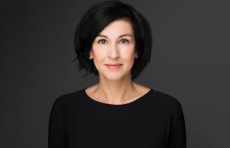 Noosha Aubel hat schwarze, schulterlange Haare. Sie trägt Lippenstift und ein schwarzes Oberteil. Sie wirkt sympathisch.