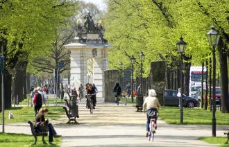 Allee im Sommer. Personen sitzen auf Bänken, fahren Fahrrad und spazieren. Im Hintergrund befindet sich das Jägertor.