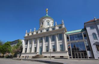 Historische Außenansicht des Potsdam Museum mit verglastem Anbau des Haupteingangs. Vor dem Gebäude spazieren zwei Personen.